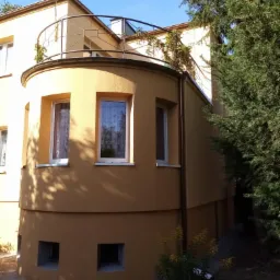 Żółta elewacja budynku z zaokrąglonym narożnikiem i balkonem z metalową balustradą, widoczna z zewnątrz w słoneczny dzień, z elementami roślinności w tle.