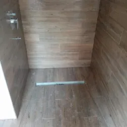 Kabina prysznicowa wyłożona brązowymi płytkami imitującymi drewno, z odpływem liniowym w podłodze i chromowaną armaturą.