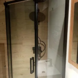 Kabina prysznicowa z czarnymi profilami i deszczownicą, widoczna umywalka i drewnopodobne płytki na ścianie.