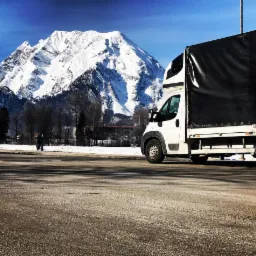 Biała furgonetka z czarną plandeką na tle ośnieżonego szczytu górskiego w słoneczny dzień. Widoczna droga i pojedyncza postać w oddali.