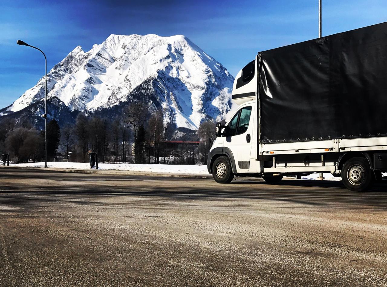Biała furgonetka z czarną plandeką na tle ośnieżonego szczytu górskiego w słoneczny dzień. Widoczna droga i pojedyncza postać w oddali.