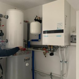 KRK SOLAR SP. Z o.o. - Instalacja kotłowni z zbiornikiem Ferolli, białym zasobnikiem i pompą ciepła Panasonic Aquarea Smart Cloud, widoczne rury, zawory i manometr, instalator podłącza przewody.