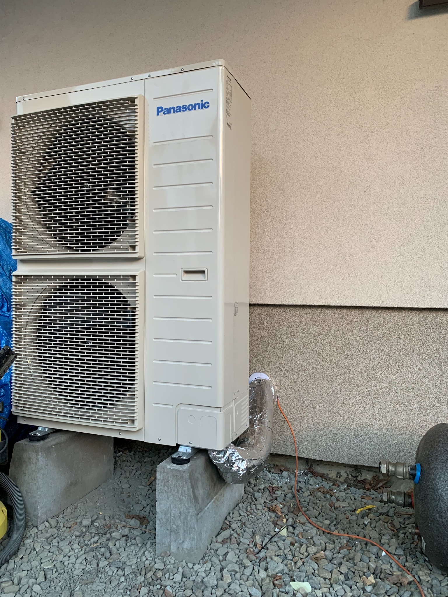 Zewnętrzna jednostka klimatyzacji Panasonic na betonowych podstawach, z widocznym przewodem w srebrnej izolacji, umieszczona na żwirowym podłożu przy jasnej ścianie budynku.