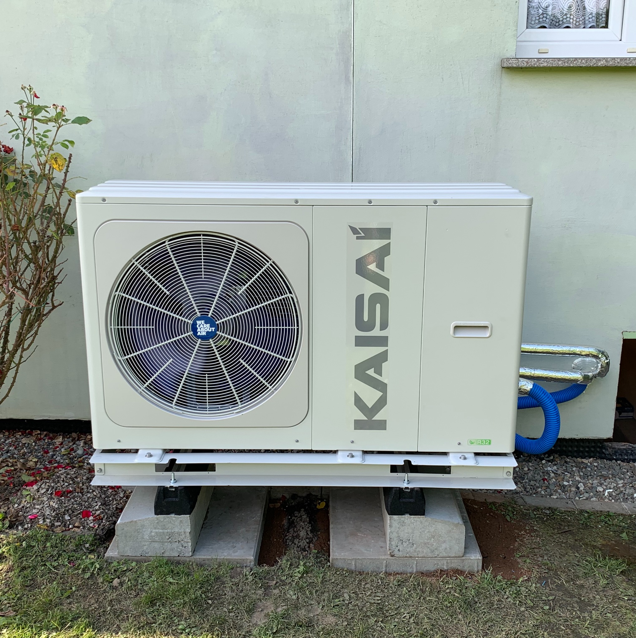Zewnętrzna jednostka pompy ciepła Kaisai zamontowana na betonowych fundamentach przy ścianie budynku z widocznymi rurami przyłączeniowymi.