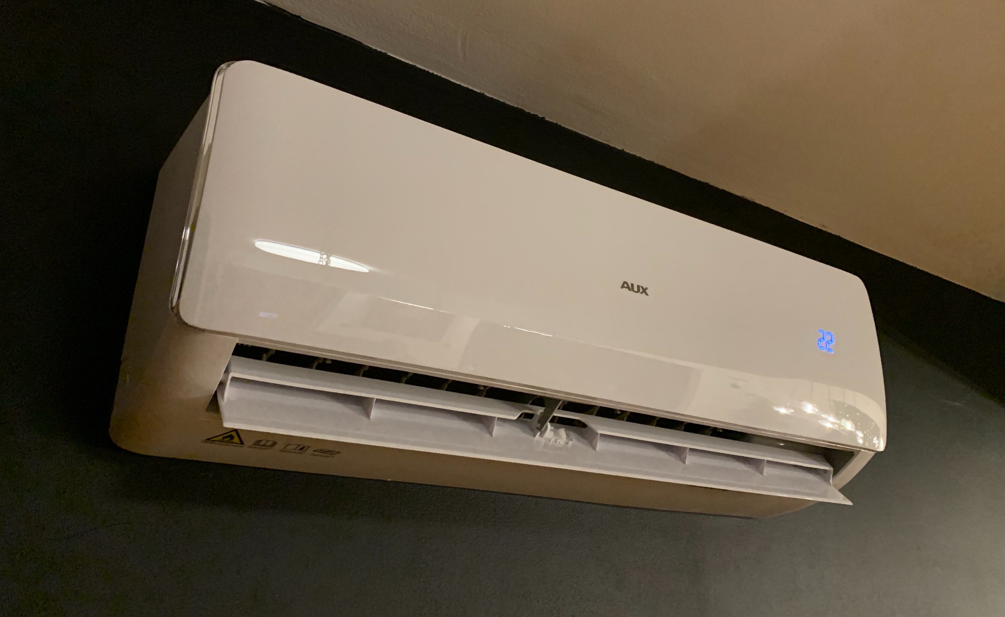 Biała jednostka wewnętrzna klimatyzacji AUX zamontowana na ciemnej ścianie, wyświetlacz pokazuje temperaturę 22 stopnie Celsjusza.