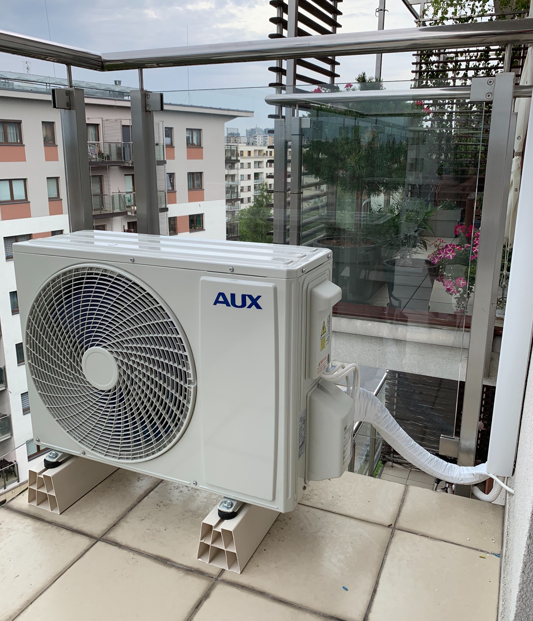 Jednostka zewnętrzna klimatyzacji AUX zamontowana na balkonie z widokiem na budynki mieszkalne, z widocznymi elementami montażowymi i izolowanym przewodem.