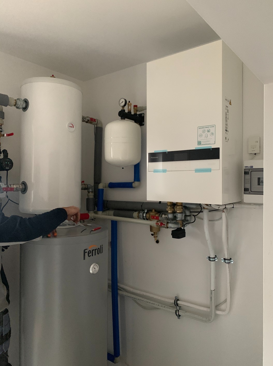Instalacja kotłowni z zbiornikiem Ferolli, białym zasobnikiem i pompą ciepła Panasonic Aquarea Smart Cloud, widoczne rury, zawory i manometr, instalator podłącza przewody.