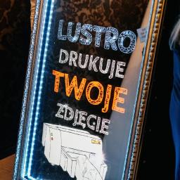 Fotoshow Sebastian Juszczyk - Podświetlane lustro z napisem 'Lustro drukuje Twoje zdjęcie' i rysunkiem drukarki, otoczone dekoracyjną ramą z niebieskim oświetleniem LED.