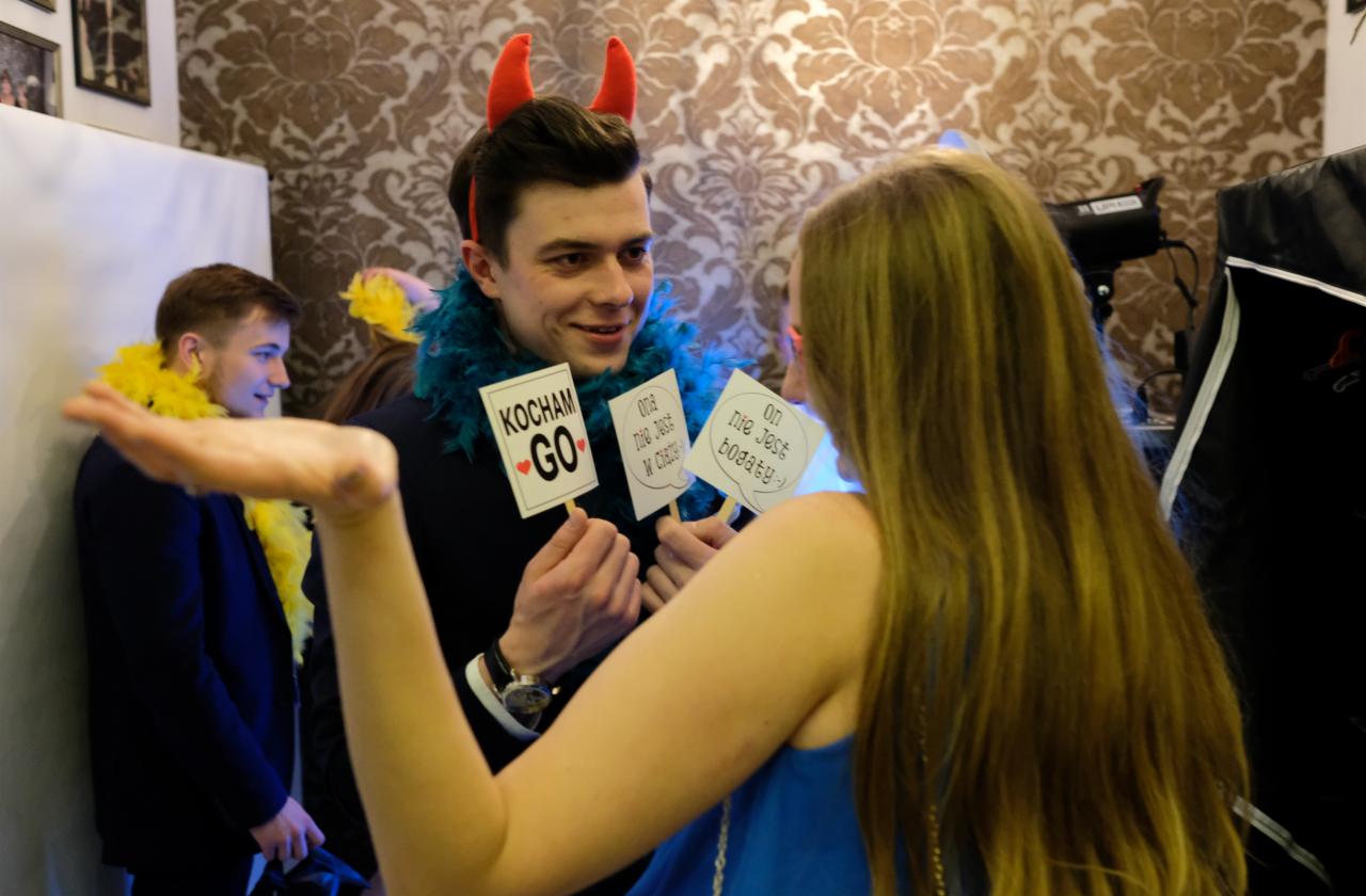 Uczestnicy zabawy w fotobudce z rekwizytami: mężczyzna z rogami diabła i niebieskim boa trzyma tabliczki z napisami 'Kocham GO' i 'Ona nie jest bogata :)' na tle wzorzystej tapety.