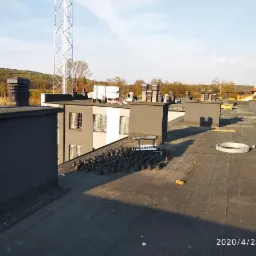 Widok z g&oacute;ry na płaski dach pokryty papą, z kominami wentylacyjnymi, elementami instalacji odgromowej i w tle budynki oraz maszt telekomunikacyjny.