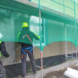 Pracownicy w kaskach na rusztowaniu, wykonujący prace na elewacji budynku, zabezpieczeni siatką ochronną.