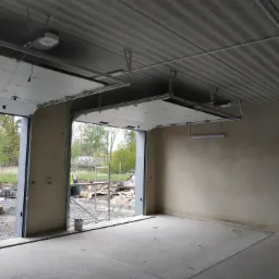 Montaż segmentowej bramy garażowej z napędem elektrycznym, widok z wnętrza surowego garażu na zewnątrz, gdzie widać rusztowanie i materiały budowlane.