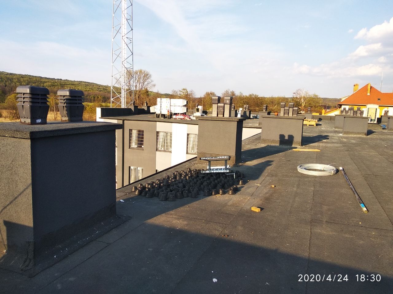 Widok z góry na płaski dach pokryty papą, z kominami wentylacyjnymi, elementami instalacji odgromowej i w tle budynki oraz maszt telekomunikacyjny.