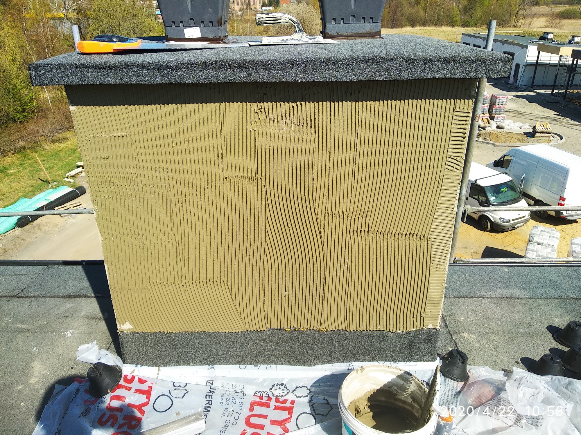 Ocieplony komin na dachu budynku w trakcie prac elewacyjnych, pokryty klejem w pionowe paski, narzędzia i wiadro z zaprawą na dachu, w tle budynki i samochody.