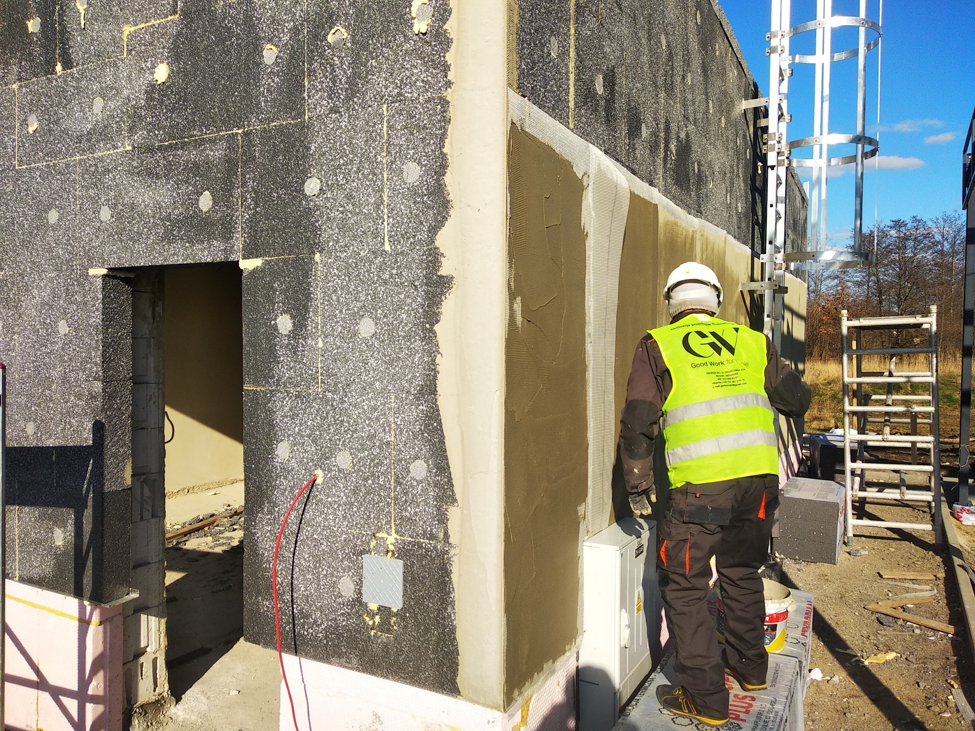 Budynek w trakcie ocieplania: szary styropian z grafitowym dodatkiem na ścianie, fragment z nałożoną siatką i klejem, pracownik w kasku i kamizelce odblaskowej z logo firmy, widoczna drabina...