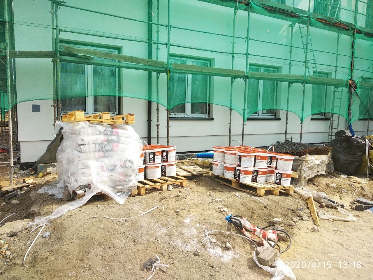 Przygotowane materiały budowlane: worki z zaprawą i wiadra z farbą na paletach, przed budynkiem w trakcie prac elewacyjnych, widoczne rusztowania z siatką ochronną.