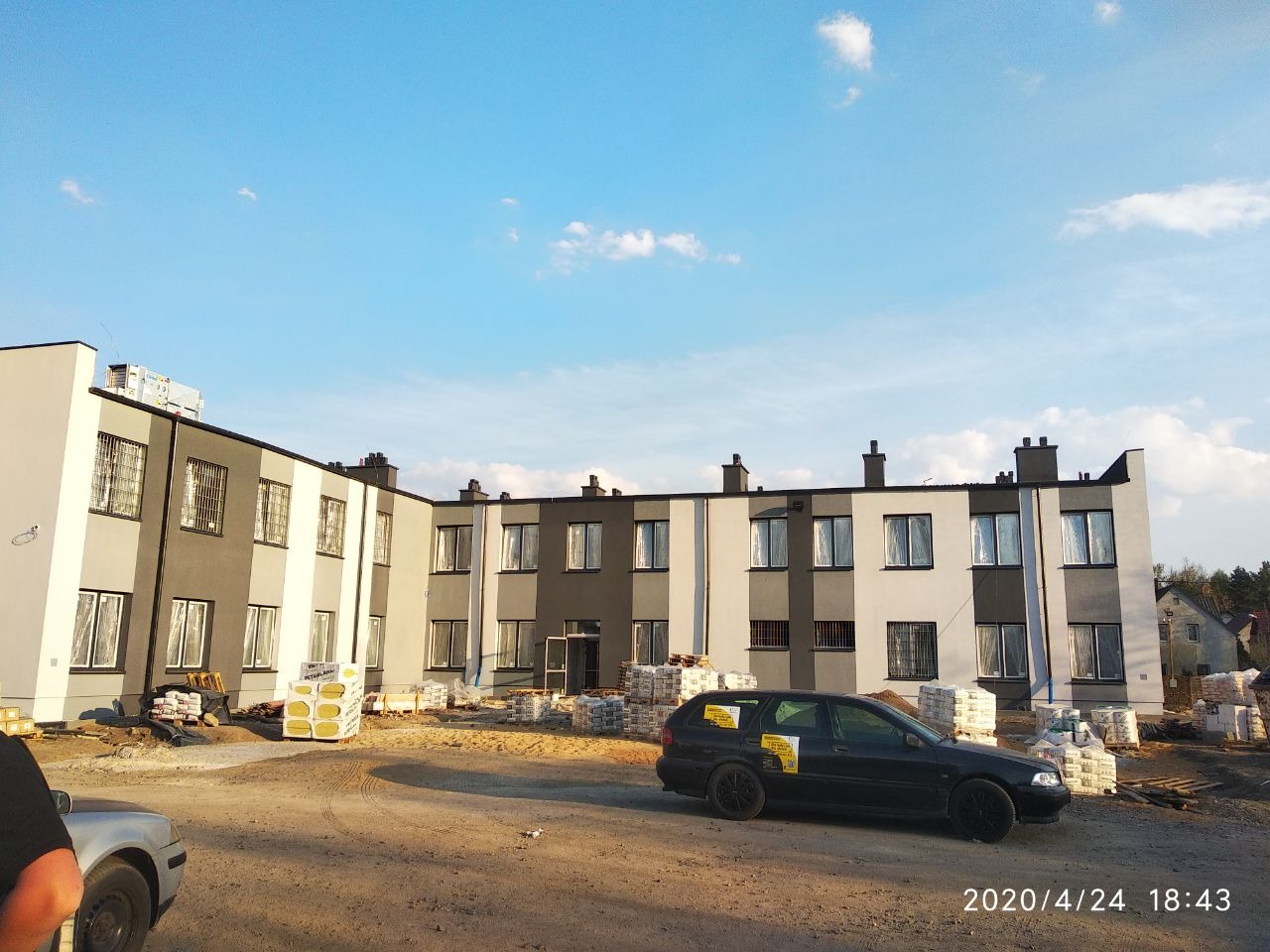 Dwukondygnacyjny budynek po remoncie elewacji w kolorach szarości i bieli, z widocznymi materiałami budowlanymi na placu budowy i zaparkowanym czarnym samochodem osobowym.