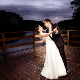 Para młoda tańczy na drewnianym pomoście nad wodą, w tle ciemny las i pochmurne niebo, delikatne oświetlenie.