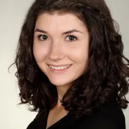 Portret uśmiechniętej kobiety z brązowymi, kręconymi włosami i piegami na twarzy, ubrana w czarną bluzkę, na jasnym tle.