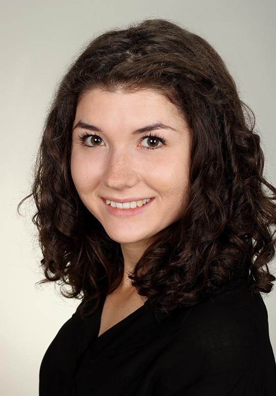 Portret uśmiechniętej kobiety z brązowymi, kręconymi włosami i piegami na twarzy, ubrana w czarną bluzkę, na jasnym tle.