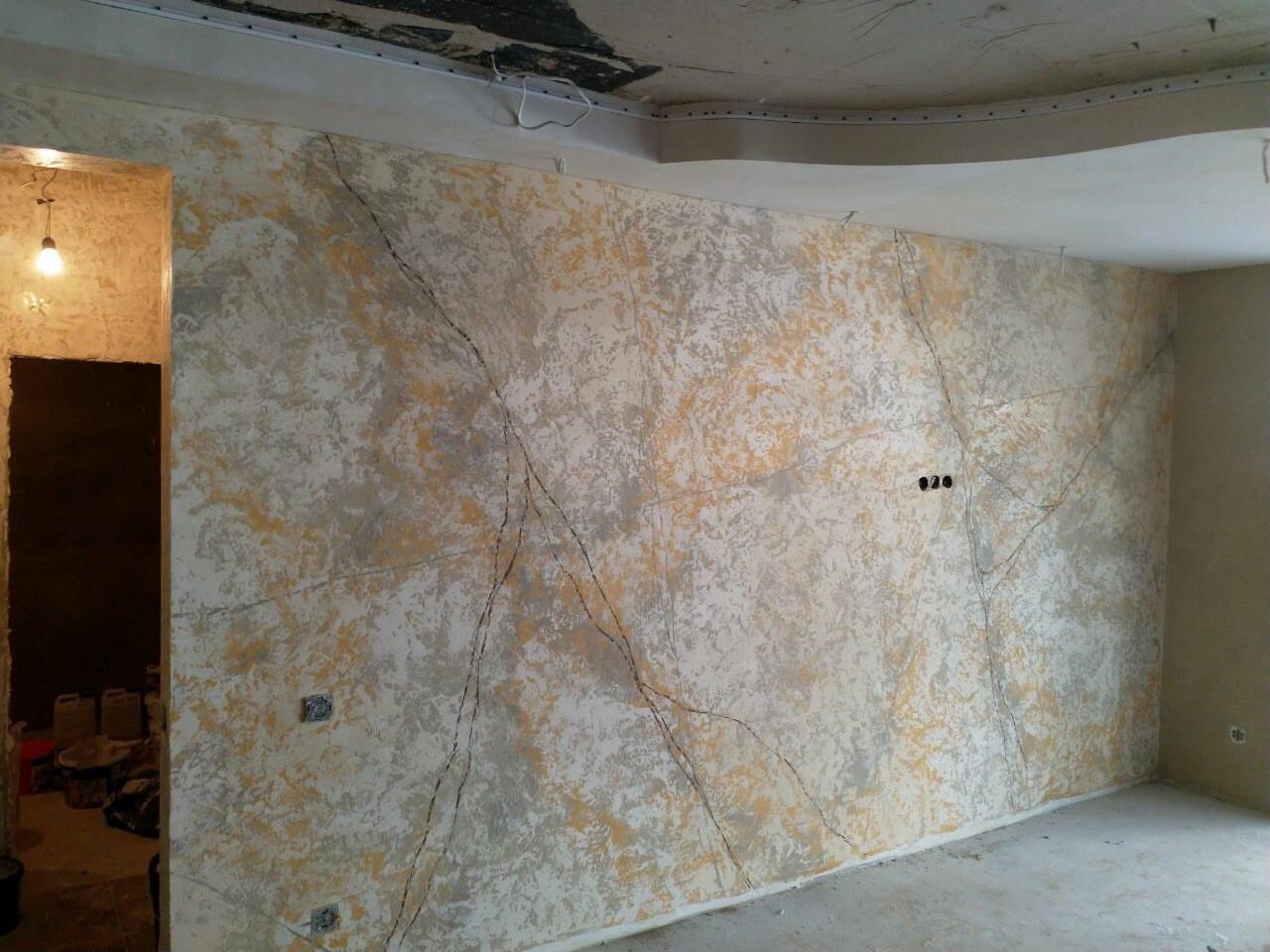 Ściana wewnętrzna z dekoracyjnym tynkiem w odcieniach bieli, szarości i żółci, wzór imitujący pęknięcia, widoczne puszki instalacyjne i surowy stan wykończenia podłogi.