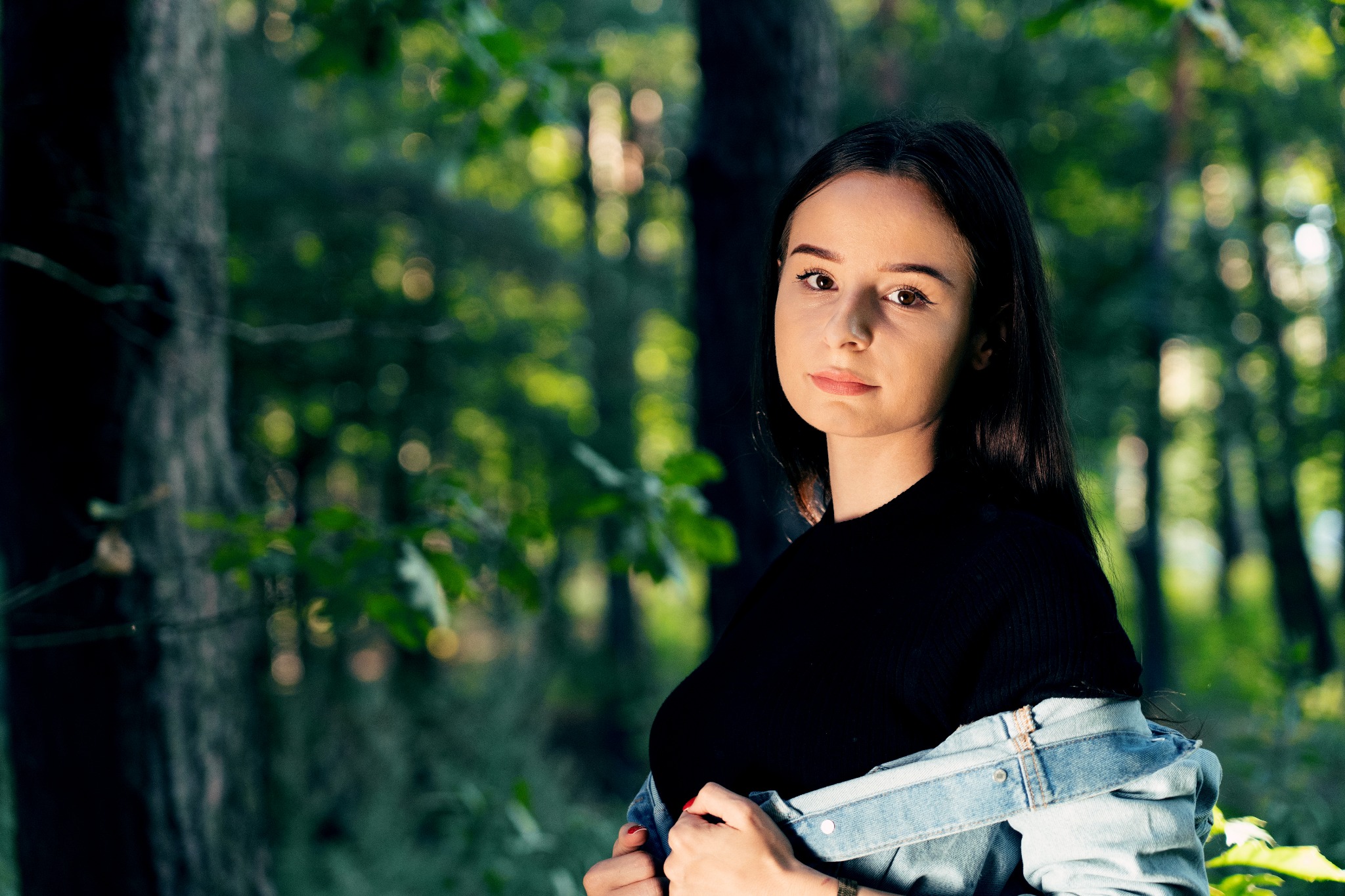 Portret młodej kobiety w lesie, ubrana w czarną bluzkę i jeansową kurtkę, z delikatnym uśmiechem i naturalnym makijażem.