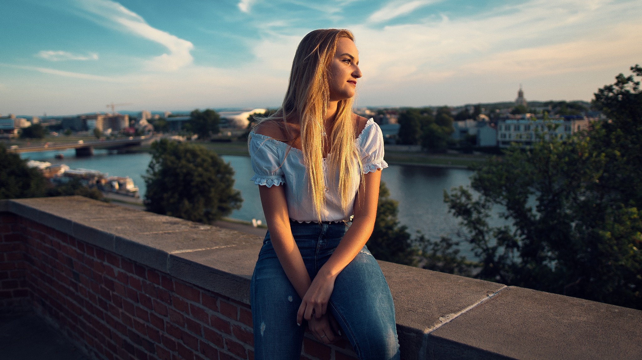 Młoda kobieta siedzi na murku z cegły, spoglądając w dal na panoramę miasta z rzeką i zielenią, w ciepłym świetle zachodzącego słońca.