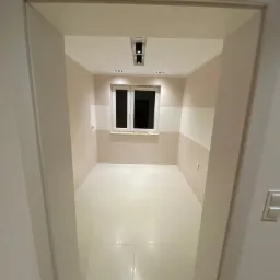 Pomalowane na dwa kolory wnętrze pokoju z białymi płytkami na podłodze, widok zza futryny drzwi, z widocznymi punktowymi lampami sufitowymi i oknem.