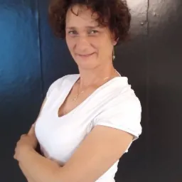 Portret uśmiechniętej kobiety z kręconymi włosami, w białej koszulce, stojącej na tle ciemnej ściany z założonymi rękami.