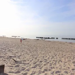 Plaża z betonowymi stopniami na pierwszym planie, w oddali osoby spacerujące brzegiem morza i drewniane pale w wodzie.