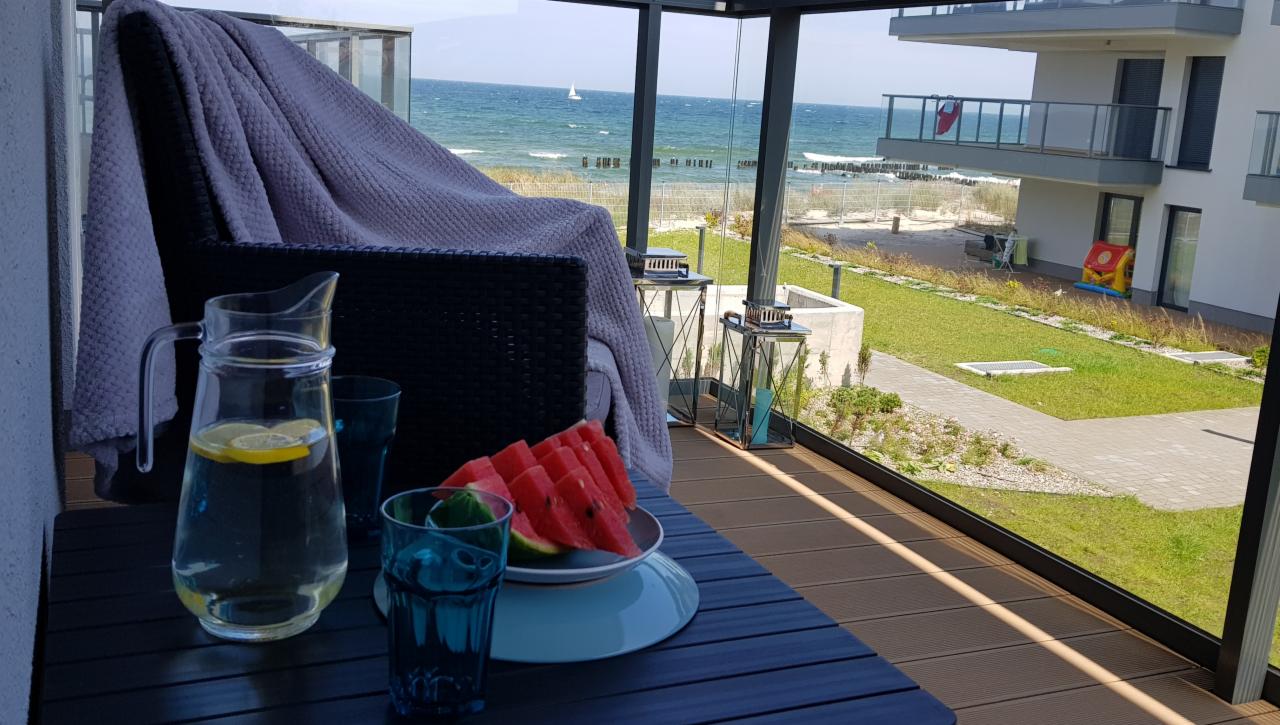 Relaksujący widok z balkonu na morze z żaglówką w tle, dzbanek z wodą z cytryną, arbuz i szklanki na stole, pleciony fotel z kocem, nowoczesne lampiony dekoracyjne, trawnik i budynek w tle.