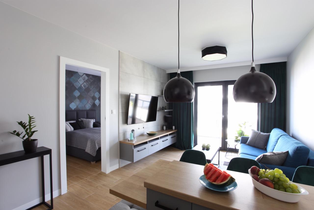 Jasne wnętrze mieszkania z otwartą przestrzenią salonu i sypialni, drewniany stół z owocami na pierwszym planie, wiszące czarne lampy, niebieska sofa i telewizor na ścianie.
