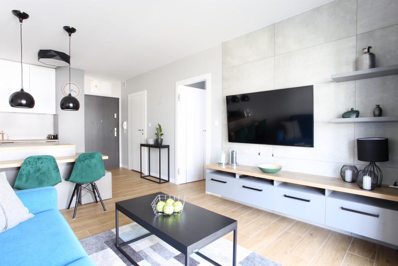 Jasne wnętrze salonu z turkusową sofą, czarnym stolikiem kawowym z jabłkami, telewizorem na ścianie z szarych paneli i drewnianą podłogą.