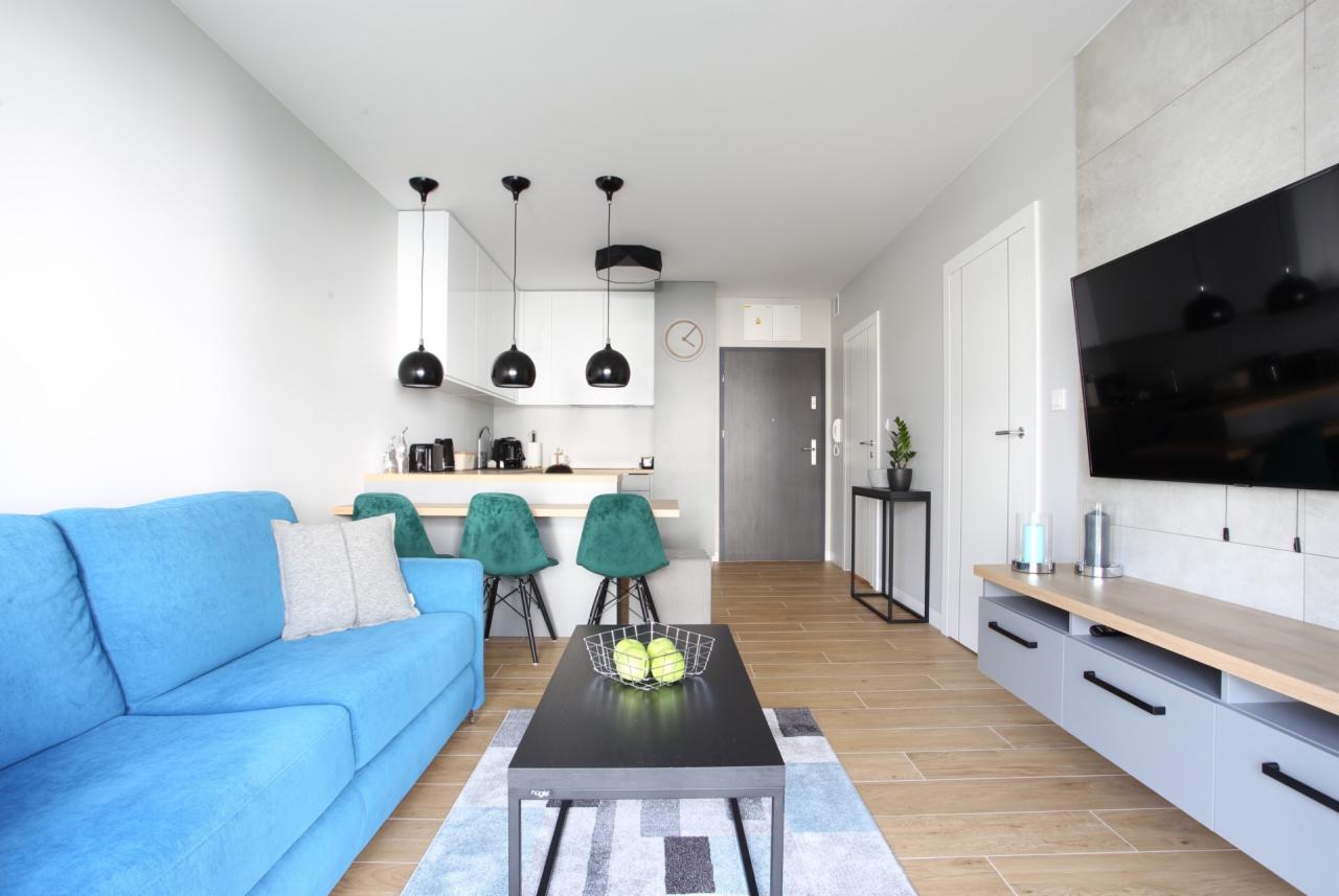 Nowoczesny salon z turkusową sofą, czarnym stolikiem kawowym z owocami, zielonymi krzesłami barowymi i aneksem kuchennym w tle.