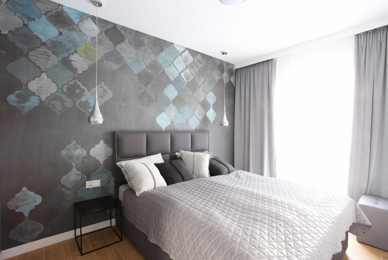 Nowoczesna sypialnia z szarą tapetą w marokańskie wzory, srebrnymi lampami wiszącymi, szarym łóżkiem z pikowaną narzutą i jasnymi poduszkami, oraz czarnym stolikiem nocnym.