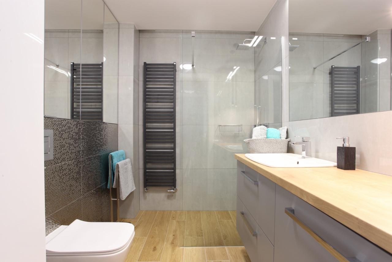 Nowoczesna łazienka z drewnianą podłogą, szarymi szafkami pod umywalką i grzejnikiem drabinkowym, widoczna kabina prysznicowa za szklaną ścianą.