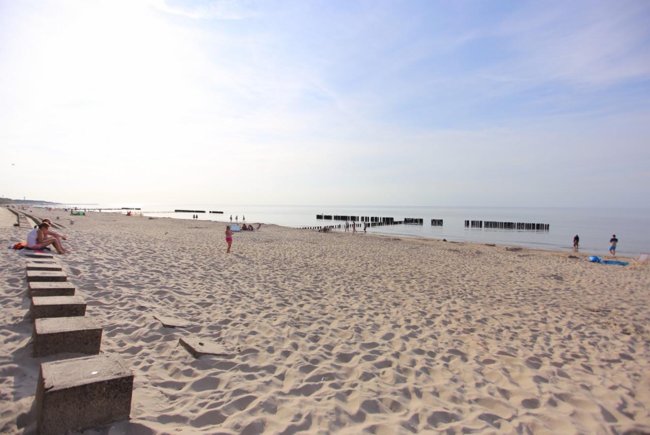 Plaża z betonowymi stopniami na pierwszym planie, w oddali osoby spacerujące brzegiem morza i drewniane pale w wodzie.