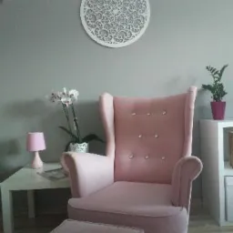 Różowy fotel uszak z podnóżkiem w Gdyni, z dekoracyjnym ażurowym panelem na ścianie i kwiatami w tle, tworzący przytulny kącik do relaksu.