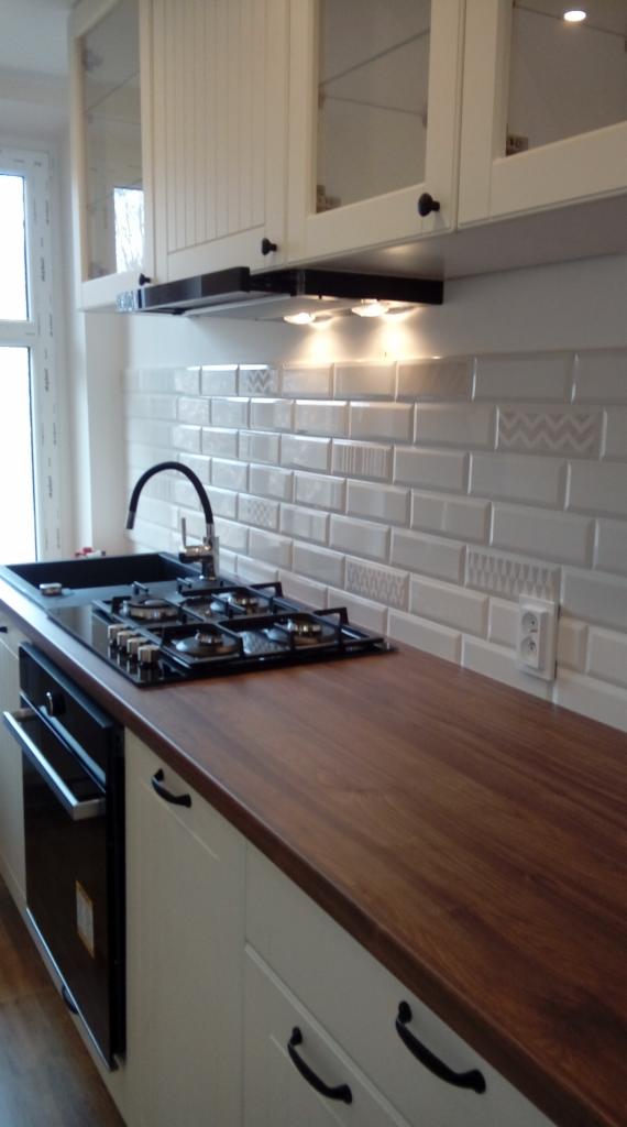 Fragment kuchni z drewnianym blatem, czarną płytą grzewczą i białymi szafkami z czarnymi uchwytami, podświetlona lampkami podszafkowymi, z dekoracyjnymi białymi płytkami na ścianie.