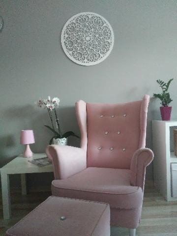 Różowy fotel uszak z podnóżkiem w Gdyni, z dekoracyjnym ażurowym panelem na ścianie i kwiatami w tle, tworzący przytulny kącik do relaksu.