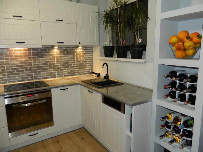 Jasna kuchnia w stylu skandynawskim z drewnianą podłogą, szarym blatem, płytkami imitującymi cegłę, oknem z roślinami w doniczkach i regałem na wino oraz misą owoców.