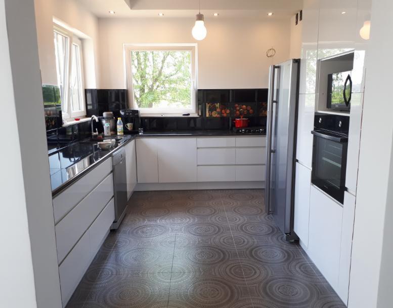 Nowoczesna, jasna kuchnia z białymi szafkami, czarnym blatem, wzorzystą podłogą i oknem z widokiem na drzewo.