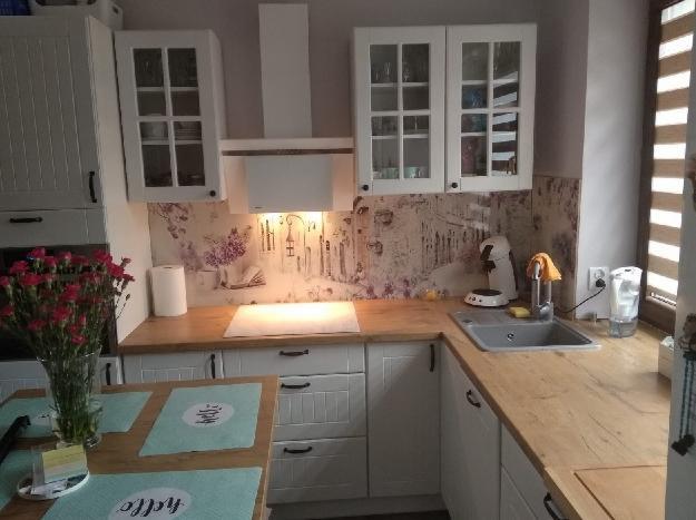 Jasna kuchnia z białymi szafkami, drewnianym blatem i dekoracyjnym panelem ściennym z motywem uliczki. Na stole wazon z czerwonymi goździkami i podkładki z napisami.