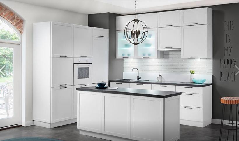 Nowoczesna, biała kuchnia z wyspą, czarnym blatem i okrągłym żyrandolem, oświetlenie podszafkowe, dekoracyjny napis na ścianie.
