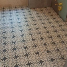 Fachowy Remont - Ułożona podłoga z płytek ceramicznych w łazience, wzór w gwiazdy i ornamenty, jasne kolory, widoczne fugi, okno w tle, ściany przygotowane pod wykończenie.