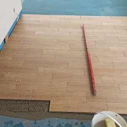 Fachowy Remont - Układanie podłogi z płytek imitujących drewno na balkonie, widoczna poziomica, klej i krzyżyki dystansowe.