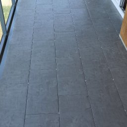 Fachowy Remont - Szare, kwadratowe płytki podłogowe na balkonie z przeszkloną balustradą i drewnianym wykończeniem ściany.