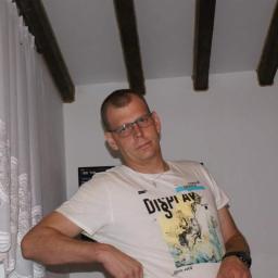 Fachowy Remont - Wnętrze pokoju z drewnianymi belkami stropowymi, koronkowymi firankami i fragmentem osoby w tle. Widoczny telewizor i białe ściany.