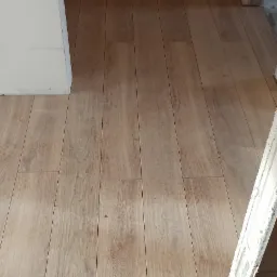 Fragment podłogi z panelami imitującymi drewno, przechodzących w jasne płytki ceramiczne przy progu drzwiowym, widoczne niedokończone wykończenie ściany po prawej stronie.