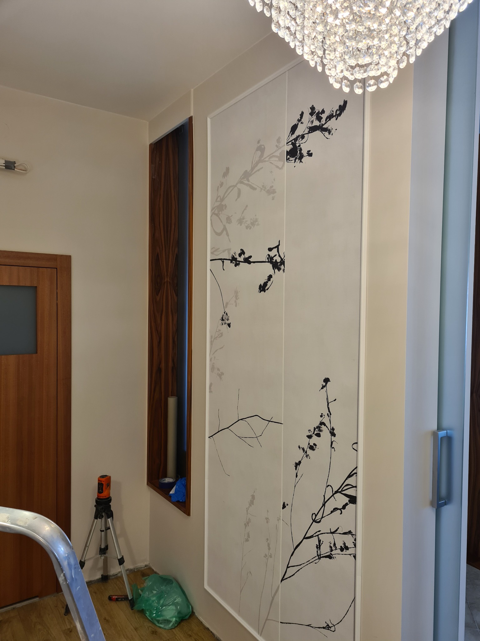 Dwa panele ścienne z motywem florystycznym, czarne gałązki na białym tle, oświetlone kryształowym żyrandolem, widoczny laser krzyżowy i fragment drabiny.